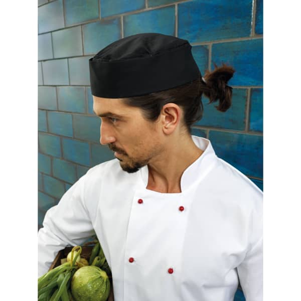 A black chef's hat is worn by a man in a white chef's coat, holding a basket of vegetables.