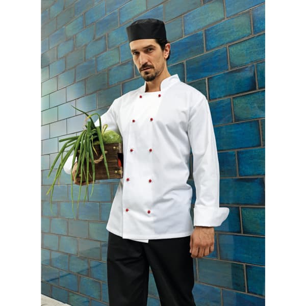 A chef wearing a white jacket and black hat, holding a wooden crate of green herbs against a blue-tiled wall.