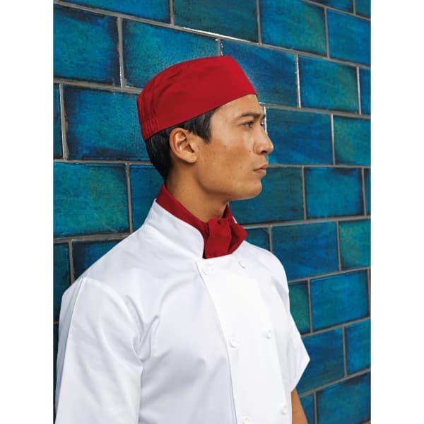 A chef wearing a red skull cap and a matching red neck scarf, standing against a blue tiled wall.
