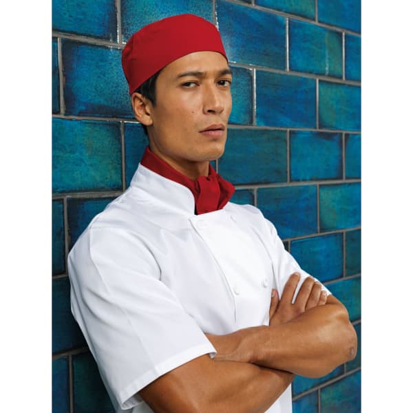 A chef wearing a red skull cap and a white uniform stands confidently against a blue tiled wall.