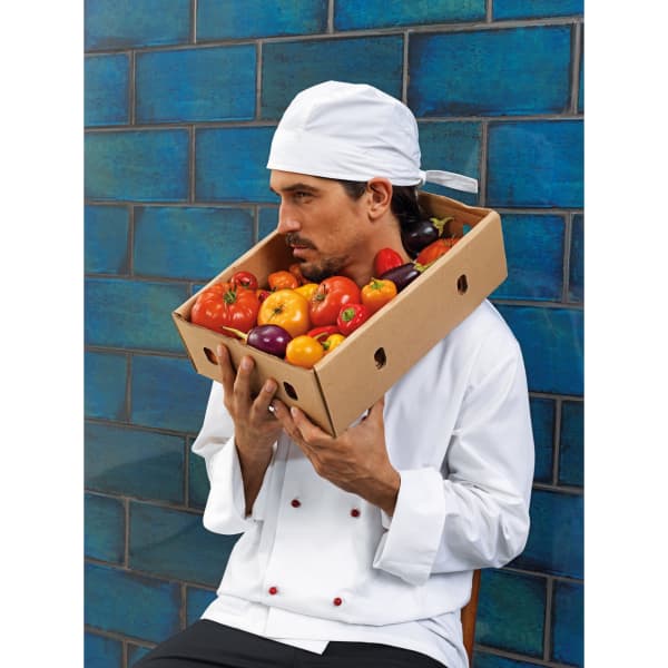 A chef in a white coat and zandana holds a cardboard box filled with colourful vegetables against a blue tiled background.