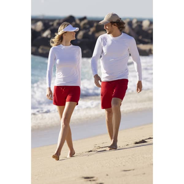 A couple walks on the beach, wearing white long sleeve rash guards and red shorts, with waves in the background.