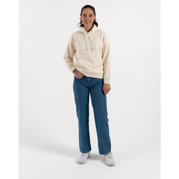 A woman in a beige hoodie and blue jeans stands on a light background, wearing white sneakers. The hoodie has a logo.