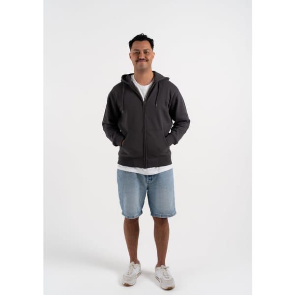 A man stands wearing a grey zip-up hoodie, paired with denim shorts and white sneakers. The hoodie has a logo.