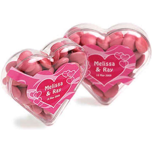 Acrylic heart container filled with pink chocolate beans, featuring a decorative heart-shaped design and a logo.
