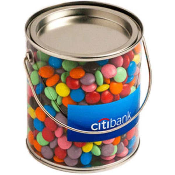 A clear PVC bucket filled with colourful choc beans, featuring a metal lid and a handle. Includes branding on the front.
