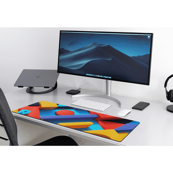 A vibrant desk mat featuring colourful geometric patterns sits on a sleek, modern desk with a computer monitor and accessories.