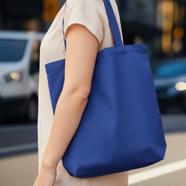 A blue reusable grocery tote bag with a single strap, held by a person on a city street.