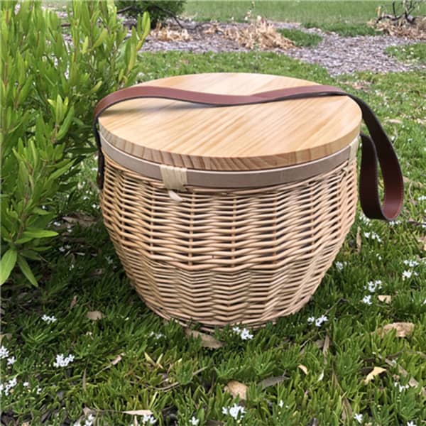 A round wicker picnic cooler basket with a wooden lid and a leather strap, set on grass with white flowers.