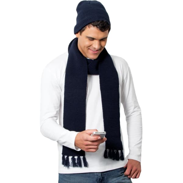 A man wearing a navy Ruga Knit Beanie and matching scarf, holding a smartphone while smiling.