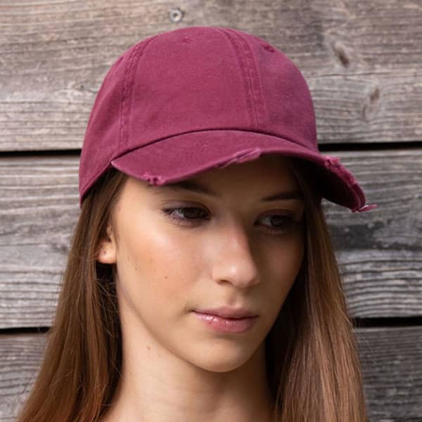 A maroon dad hat with a slightly distressed design, featuring a logo, displayed on a model in front of wooden background.