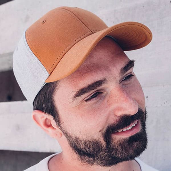 A cap featuring a brown leather front and grey fabric sides, worn by a man with a beard against a concrete background.