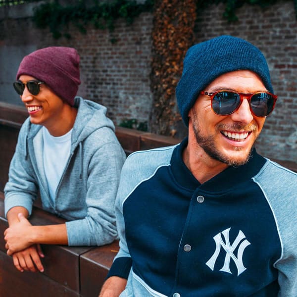 Two men wearing beanies, one in dark blue and the other in maroon, along with sunglasses, sitting outdoors.