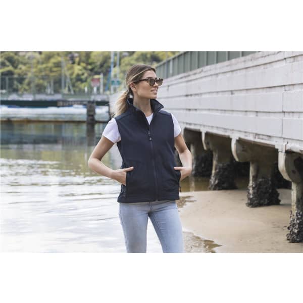 A woman wearing a black softshell vest over a white T-shirt, standing near the water with hands in pockets.