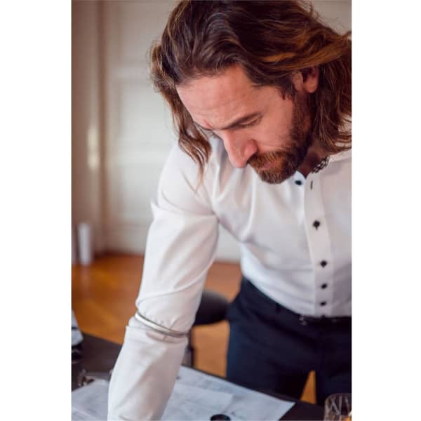 A man in a white button-up shirt with black buttons leans over a table, working on documents.