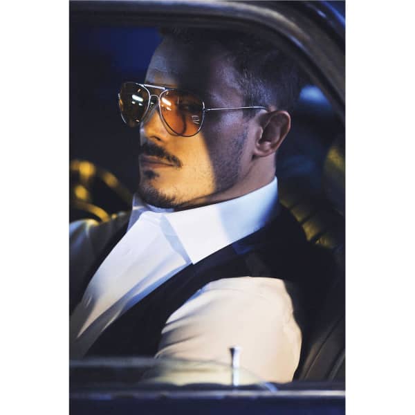 A man in a car wearing sunglasses and a black vest over a white button-up shirt.