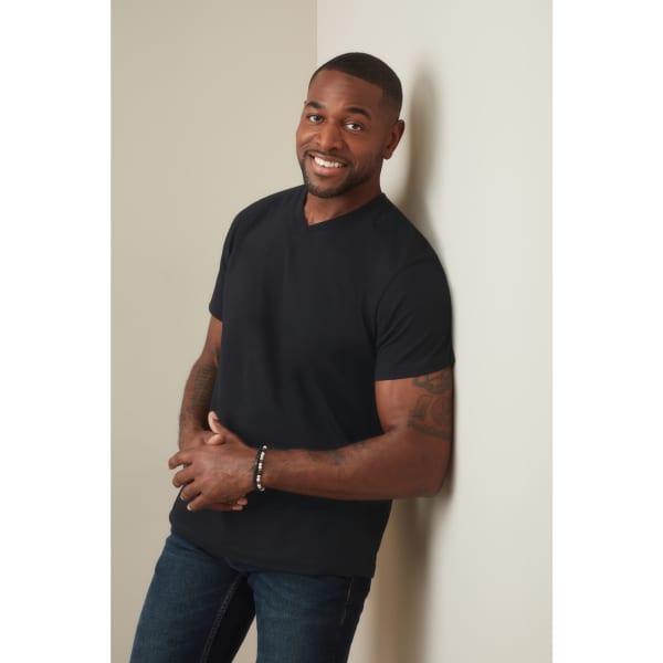 A man wearing a black classic V-neck T-shirt, standing against a wall with a friendly smile.