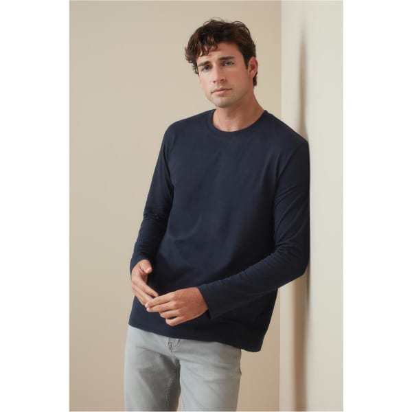A man models a navy long sleeve T-shirt, leaning against a beige wall, styled with light-coloured jeans.