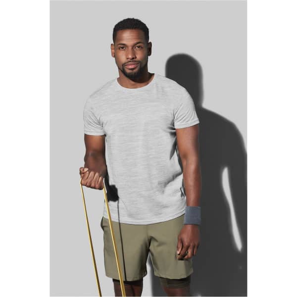 A man wearing a grey athletic T-shirt and olive shorts, holding a skipping rope, stands against a grey background.