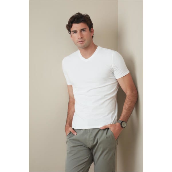 A man in a white V-neck t-shirt stands against a neutral background, hands in pockets, wearing casual pants.