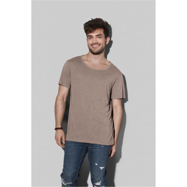 A light brown crew neck t-shirt worn by a smiling man, paired with denim jeans, against a grey background.