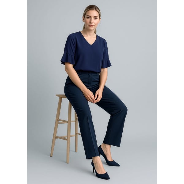 A woman sits on a stool wearing navy slacks and a matching top, styled with heels, against a grey backdrop.