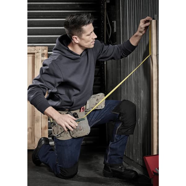 A kneeling man measuring a wooden board, wearing a grey hoodie and a tool belt, with dark pants and shoes.