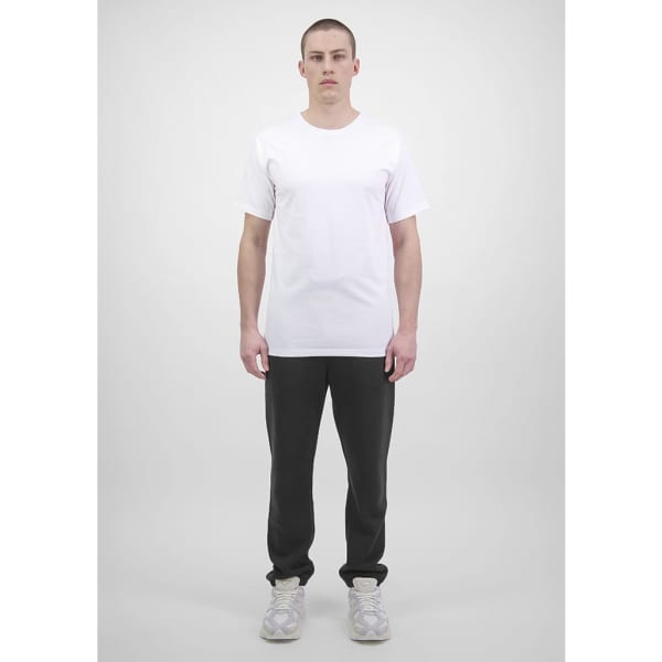 A man stands wearing a plain white t-shirt and black track pants. The track pants feature a logo.