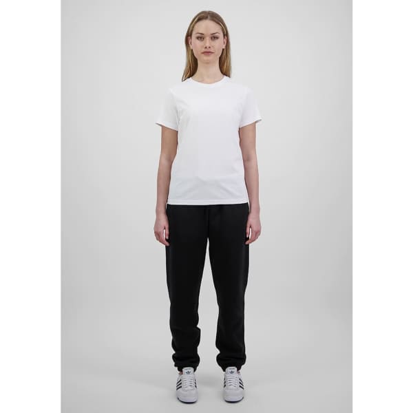 A woman in a white t-shirt and black track pants stands against a plain background. The pants have a branding logo.