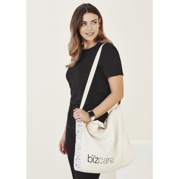 A woman wearing a black top carries a cream unisex tote bag with a logo, featuring a simple design.