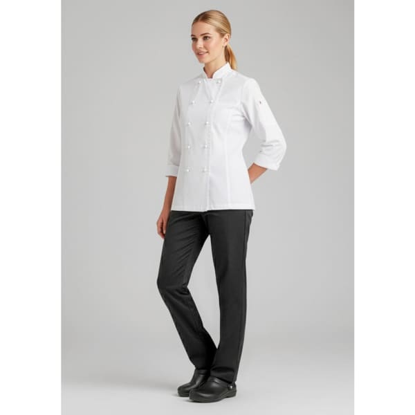 A woman in a white chef's jacket and black pants stands confidently against a neutral background. The attire includes a logo.