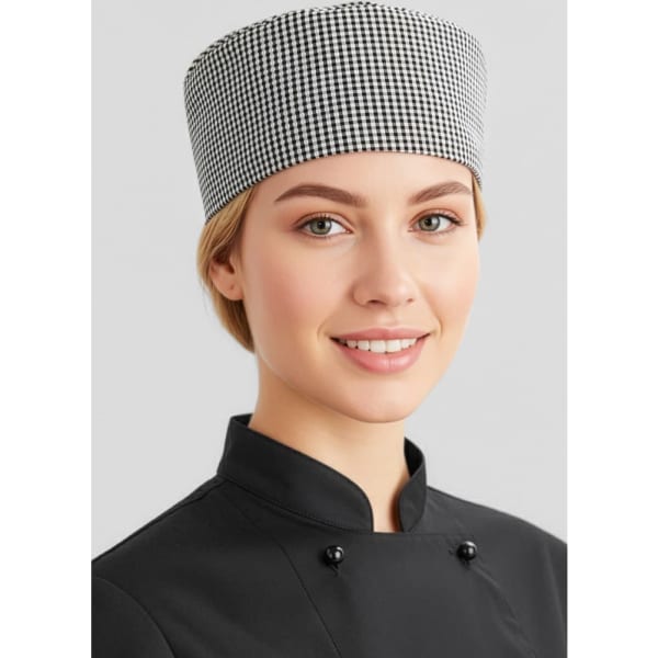 A chef cap in a black and white checkered pattern worn by a smiling individual in a black chef uniform.