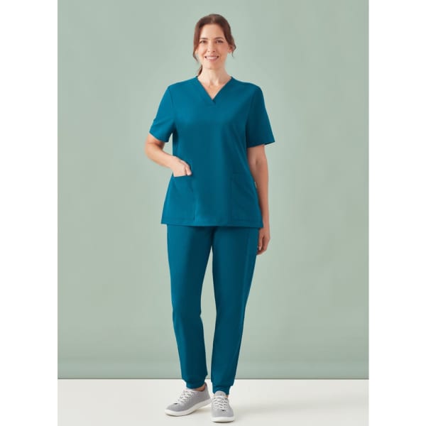 A woman stands wearing teal jogger scrub pants and a matching scrub top, with a pocket on the top and a logo visible.