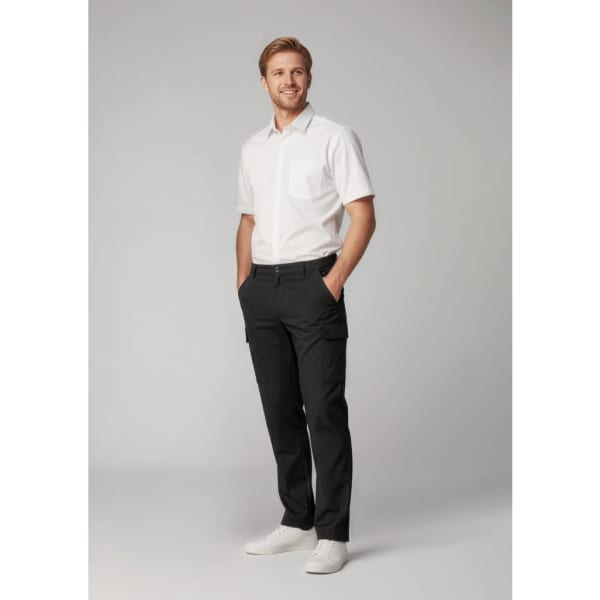 A man stands wearing black cargo pants and a short-sleeve white shirt, with hands in pockets and a smile.