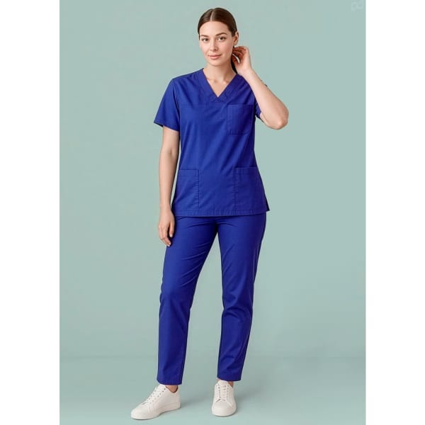 A woman in a blue scrub top and matching scrub pants stands against a green background, wearing white shoes.