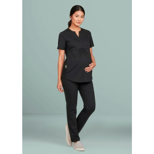 A woman in black maternity scrub pants stands against a light green background, wearing a black scrub top.