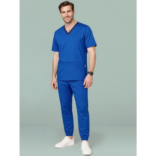A man in a blue scrub top and slim leg jogger scrub pants, wearing white sneakers, standing against a green background.