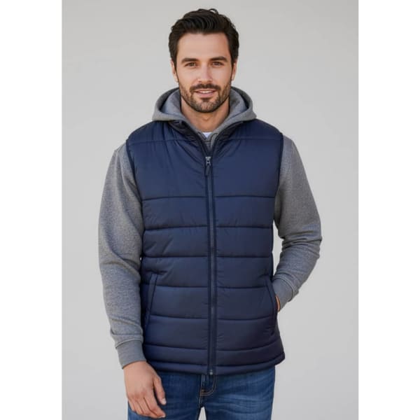 A navy blue men's alpine vest with a padded design, worn over a grey hoodie. The model is smiling and standing casually.