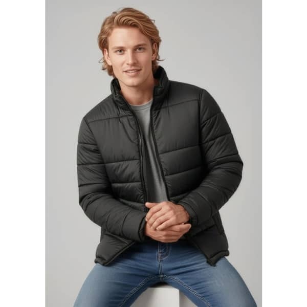 A puffer jacket in black, featuring a zip front and standing collar, worn by a model seated on a white surface.