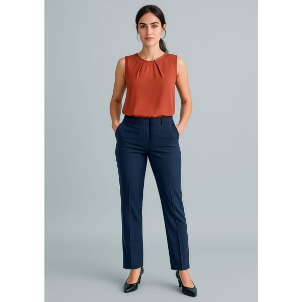 A woman in a sleeveless rust top and navy tapered leg pants, standing with hands in pockets, wearing heels.