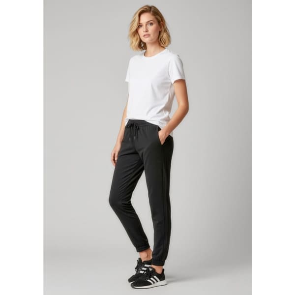 A woman stands wearing black track pants with a drawstring and a white t-shirt, featuring a logo on the pants.