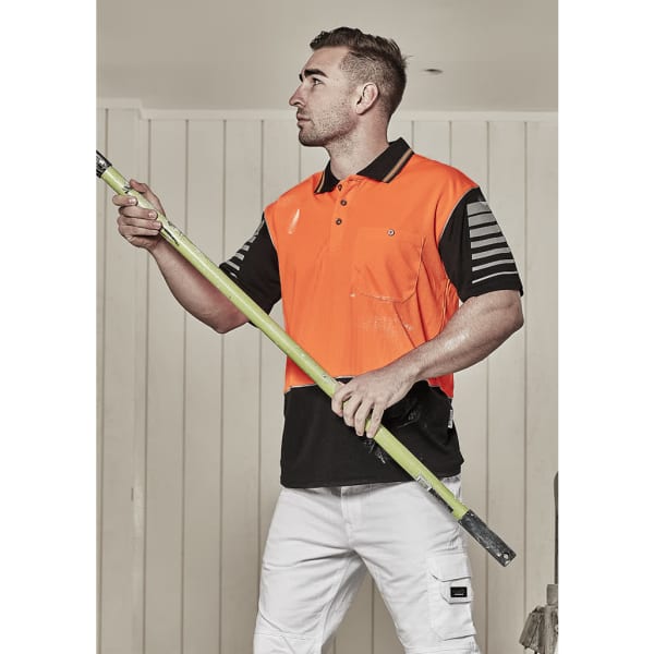 A man wearing an orange and black short sleeve polo shirt holds a green pole, standing against a light-coloured background.