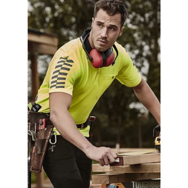 A man wears a high-visibility yellow short sleeve polo shirt with black stripes, featuring a logo. He is outdoors, working.