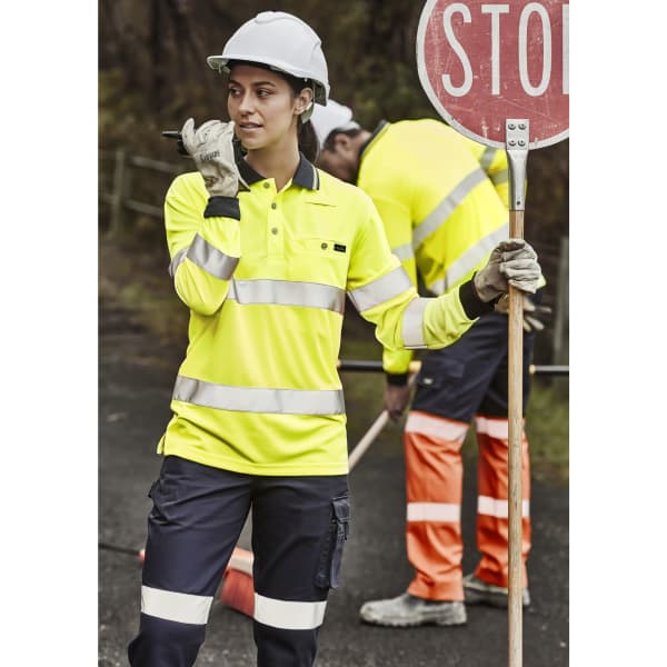 A woman in a high-visibility yellow polo shirt with reflective tape, wearing a hard hat and gloves, holds a sign.