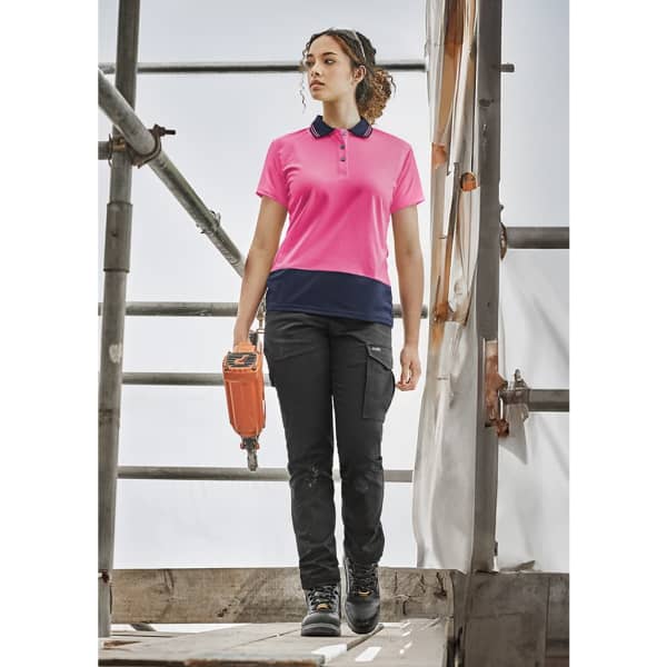 A woman wearing a pink and navy short sleeve polo shirt stands on scaffolding, holding a tool. The shirt features a logo.