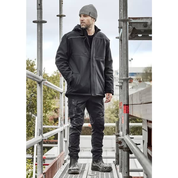 A man in a black unisex softshell jacket stands on scaffolding, wearing black pants and a beanie. The jacket has a logo.