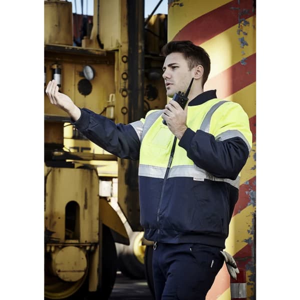 A man in a high visibility quilted flying jacket, featuring yellow and navy colours, is using a walkie-talkie.