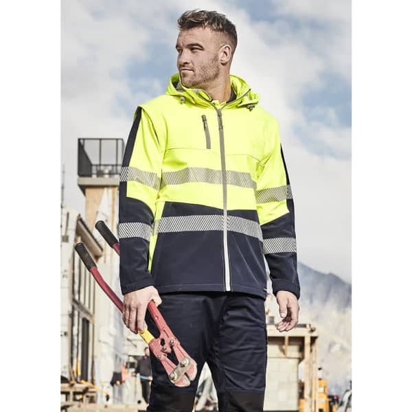 A high-visibility unisex softshell jacket in yellow and navy, featuring reflective stripes, worn by a man holding tools.