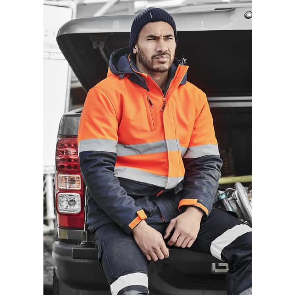 A hi-vis orange and navy softshell jacket, featuring reflective stripes, worn by a man seated on a vehicle's tailgate.
