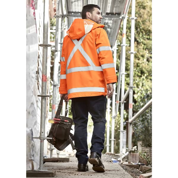 A man wearing an orange waterproof jacket with reflective stripes and carrying a tool bag walks on a construction site.